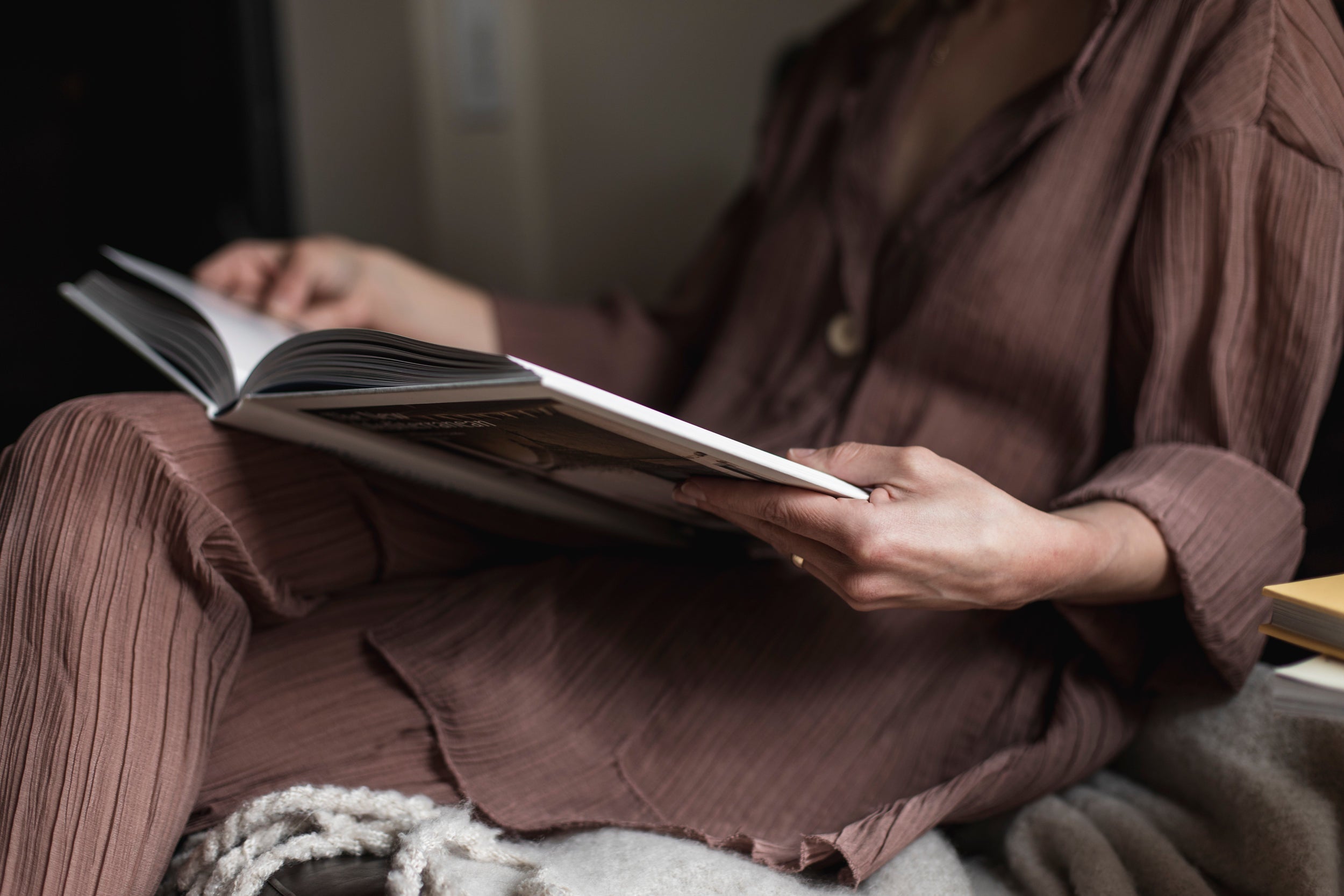 Person sitzt entspannt und liest ein aufgeschlagenes Buch in gemütlicher Atmosphäre. Das warme Licht und die weiche Kleidung schaffen ein ruhiges, behagliches Ambiente
