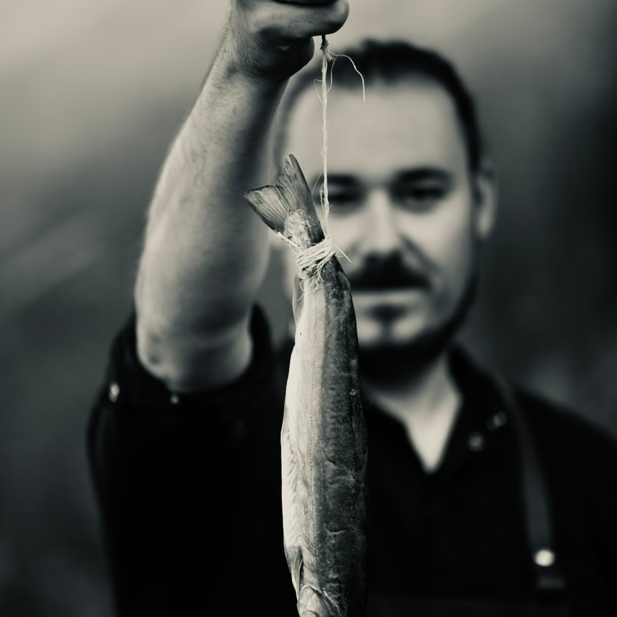 Porträtfoto von Christoph Krabichler. Der Autor hält einen Fisch an einer Schnur in die Kamera, der Fokus liegt auf dem Fisch im Vordergrund, während Krabichler unscharf im Hintergrund zu sehen ist. Schwarz-Weiß-Aufnahme.