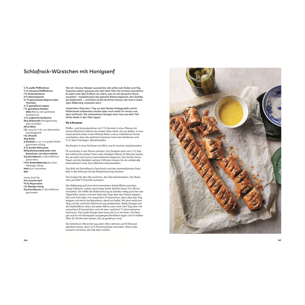 Schlafrock-Würstchen mit Honigsenf: Zwei goldbraun gebackene Blätterteigrollen, gefüllt mit Würstchen, angeschnitten auf Backpapier mit Schälchen Honigsenf-Dip, eingelegten Gurken und buntem Geschirr.