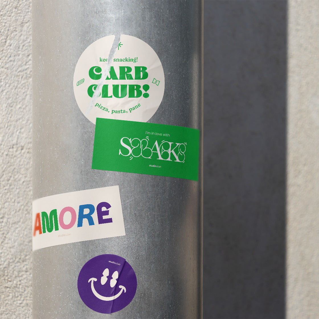 Mehrere AMORE Sticker von studio ciao auf einem Metallpfosten im urbanen Umfeld, darunter Motive wie „Carb Club“, „Snacks“, „AMORE“ und ein Smiley-Sticker, aufgeklebt im Streetstyle-Look bei Tageslicht.