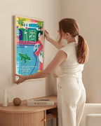 Fine Art Print French Riviera von Jean Philippe Henric in hellem Holzrahmen an der Wand. Eine Frau hängt den farbenfrohen Kunstdruck mit Poolszene, Krokodil und Palmen auf. Zeitgenössische Kunst mit mediterranem Flair für moderne Wohnräume.