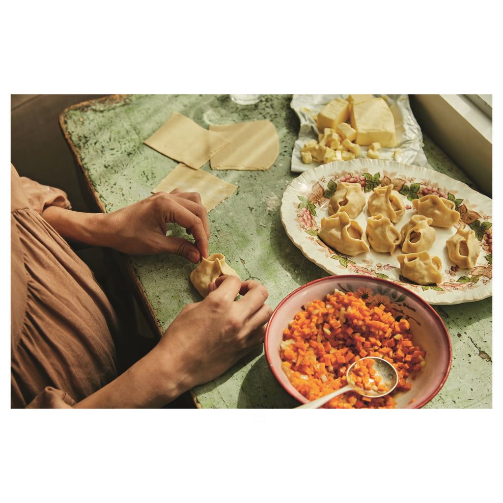 Zubereitung von Teigtaschen auf einem grünen Holztisch. Hände formen gefüllte Teigtaschen, daneben Schüssel mit gewürfelten Karotten und Teller mit fertigen Teigtaschen.