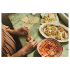 Zubereitung von Teigtaschen auf einem grünen Holztisch. Hände formen gefüllte Teigtaschen, daneben Schüssel mit gewürfelten Karotten und Teller mit fertigen Teigtaschen.