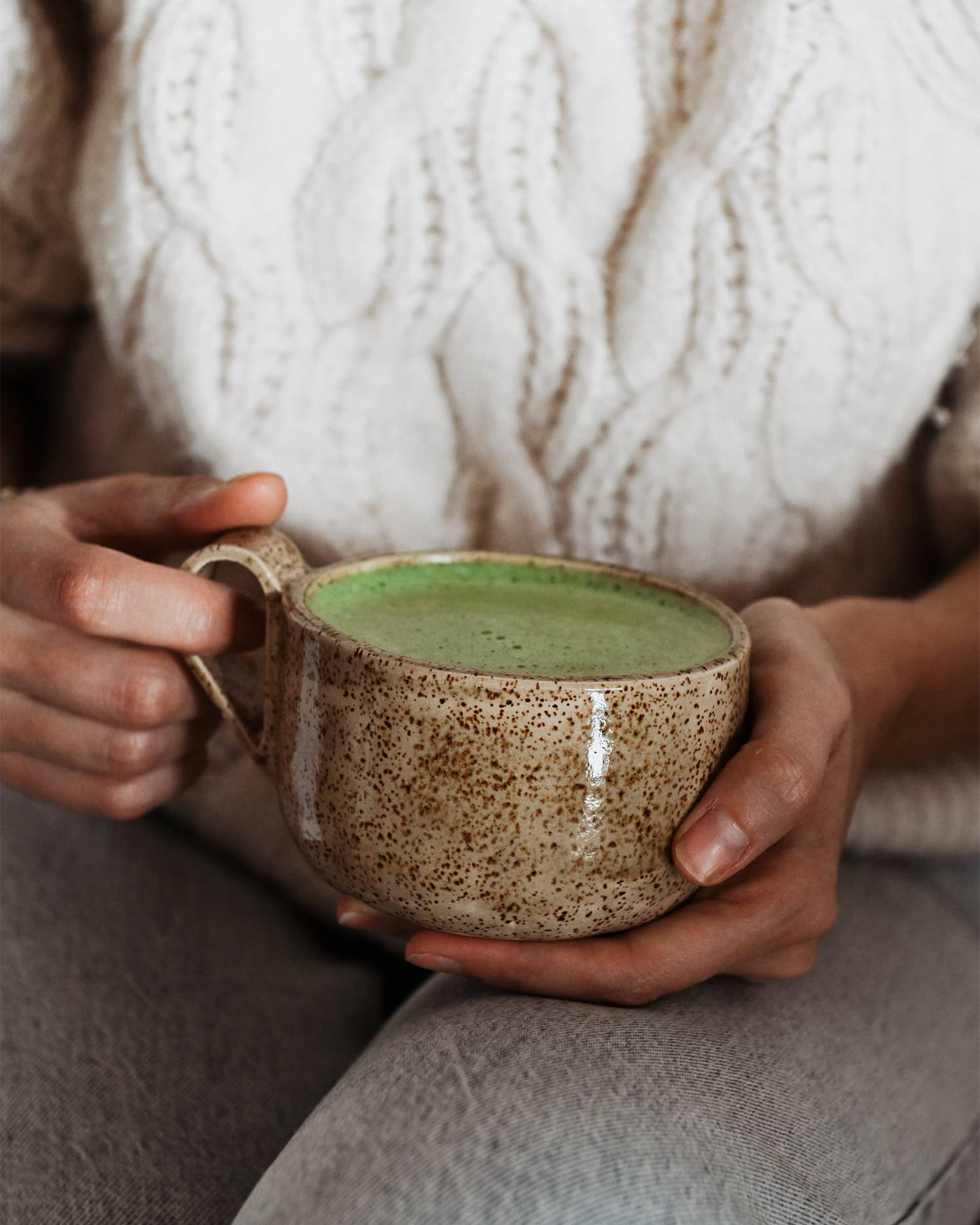 Handgefertigte Keramiktasse Caliente von Üm Studio in warmem Beige mit feinen braunen Sprenkeln, gehalten in beiden Händen. Gefüllt mit cremigem Matcha Latte, ein Moment der Ruhe und Achtsamkeit, der die Wärme Málagas in den Alltag bringt.