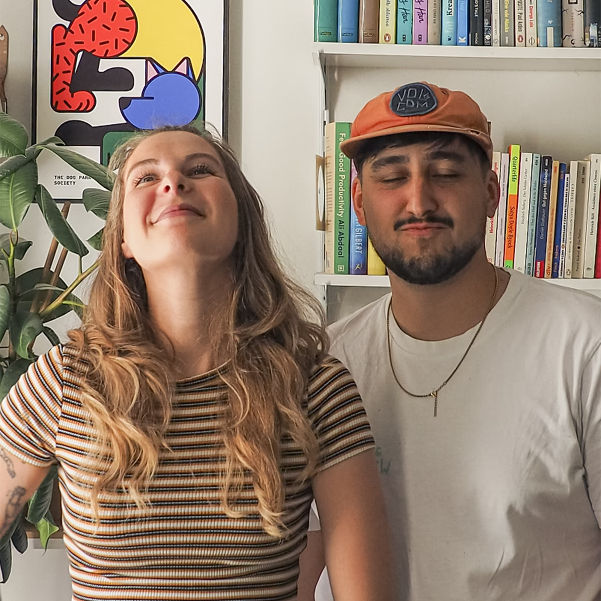 Porträtfoto des Design-Duos RadandYellow aus Wien, bestehend aus Lisa und Johann. Beide stehen lächelnd vor einem Bücherregal, sie trägt ein gestreiftes Shirt, er eine orangefarbene Cap und ein weißes T-shirt mit einer goldene Kette.