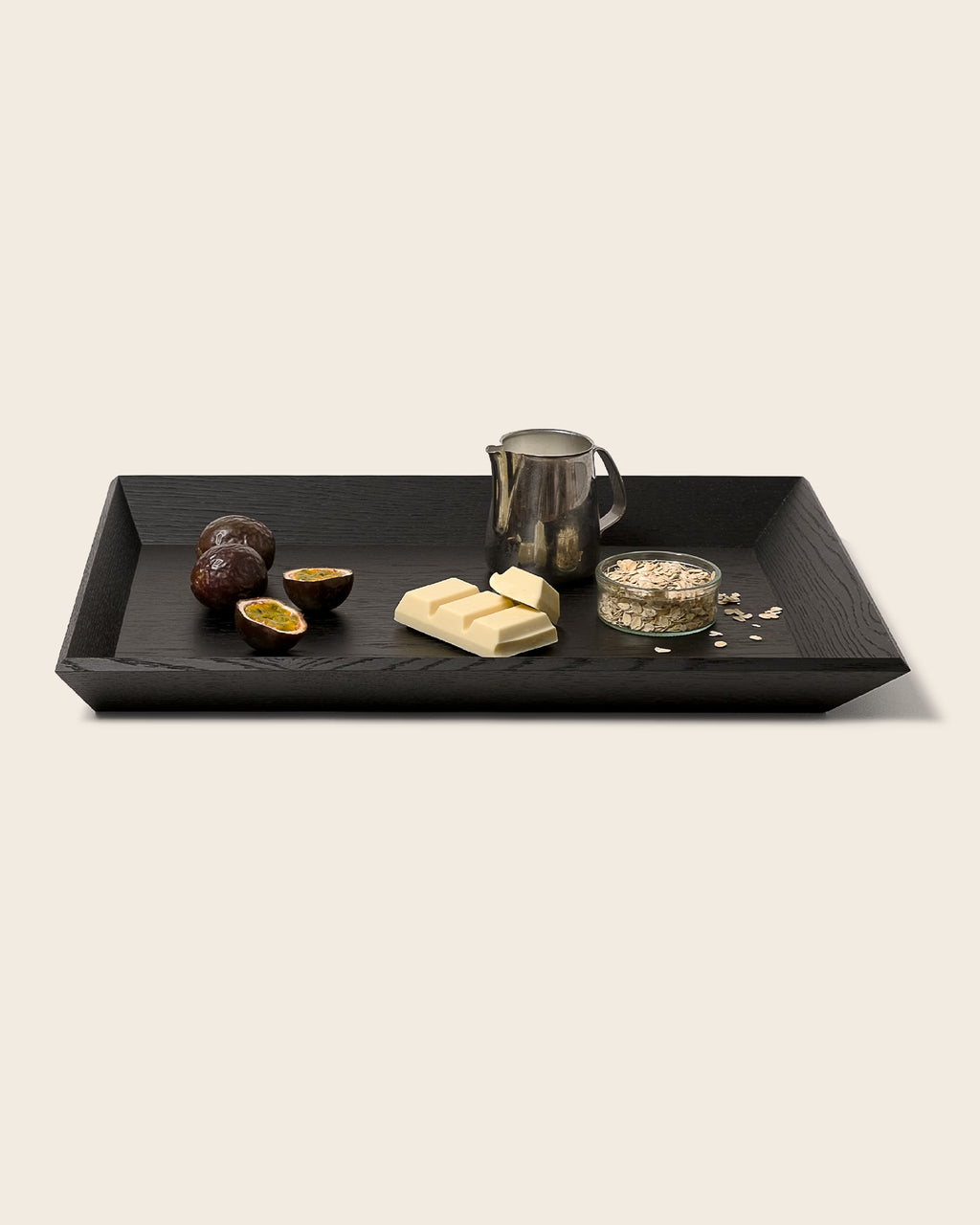 Schwarzes Holztablett OBLIQUE von NIEVEN mit schrägen Kanten. Darauf arrangiert: Passionsfrüchte, weiße Schokolade, eine Milchkanne und ein Glas Haferflocken. Modernes, minimalistisches Serviertablett im architektonischen Design von Jörg Adam.