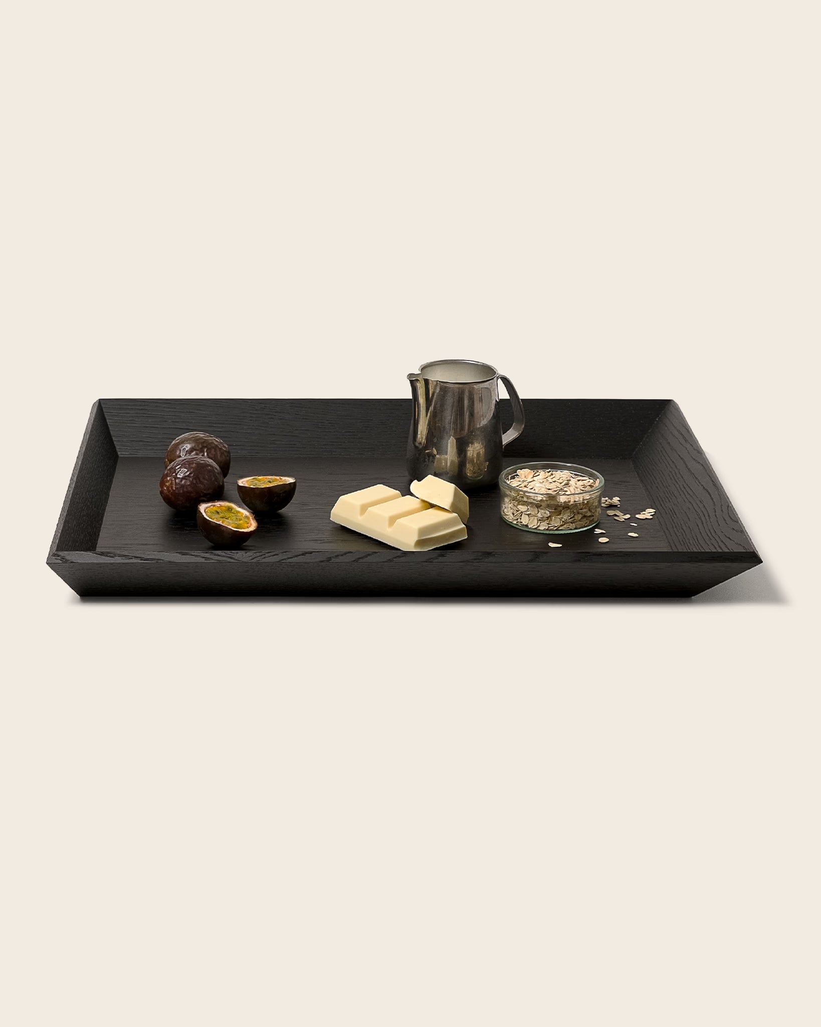 Schwarzes Holztablett OBLIQUE von NIEVEN mit schrägen Kanten. Darauf arrangiert: Passionsfrüchte, weiße Schokolade, eine Milchkanne und ein Glas Haferflocken. Modernes, minimalistisches Serviertablett im architektonischen Design von Jörg Adam.