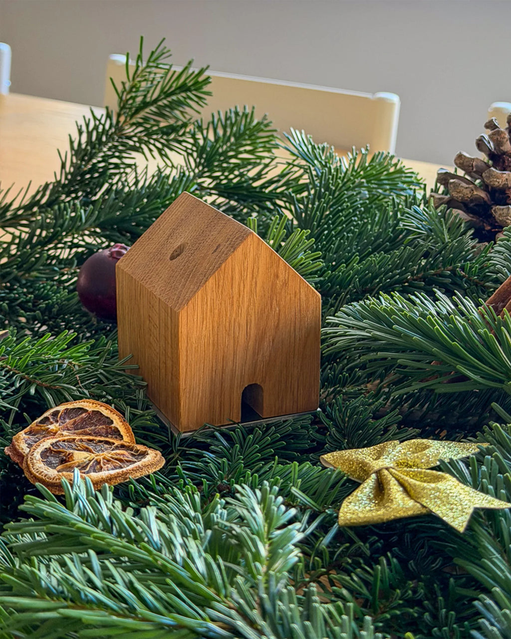 Handgefertigtes Räucherhäuschen Wieden aus recyceltem Wiener Eichenholz auf Edelstahlplatte. Platziert auf grünen Tannenzweigen und umgeben von Zimt, einer goldenen Schleife, einem Apfel, Orangenschreiben und einem Tannenzapfen.