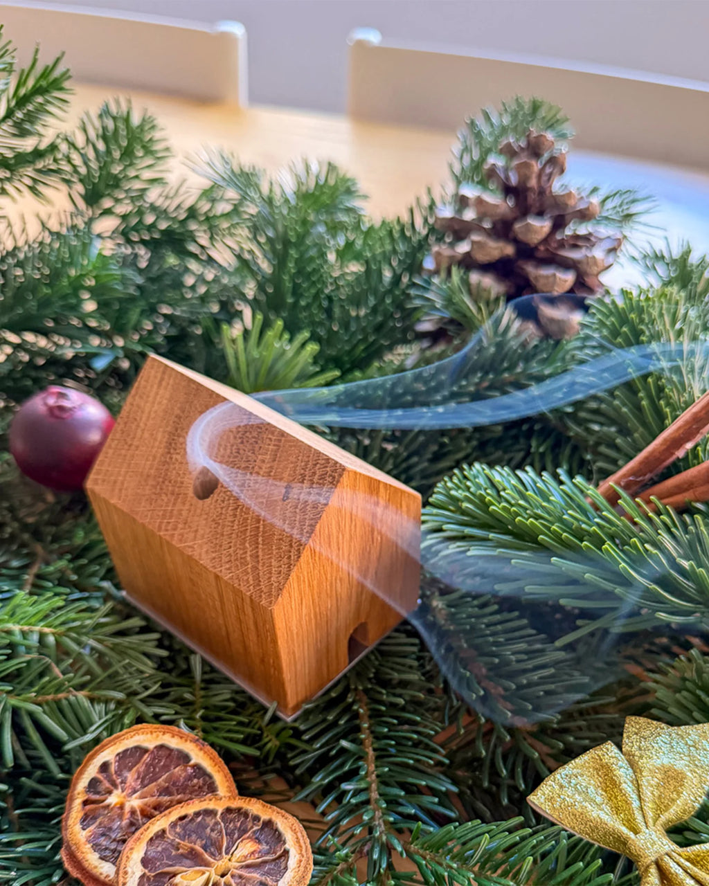 Handgefertigtes Räucherhäuschen Wieden aus recyceltem Wiener Eichenholz auf einer Edelstahlplatte. Das Räucherhäuschen raucht. Platziert auf grünen Tannenzweigen und umgeben von Zimt, einer goldenen Schleife, einem Apfel, Orangenschreiben und einem Tannenzapfen.