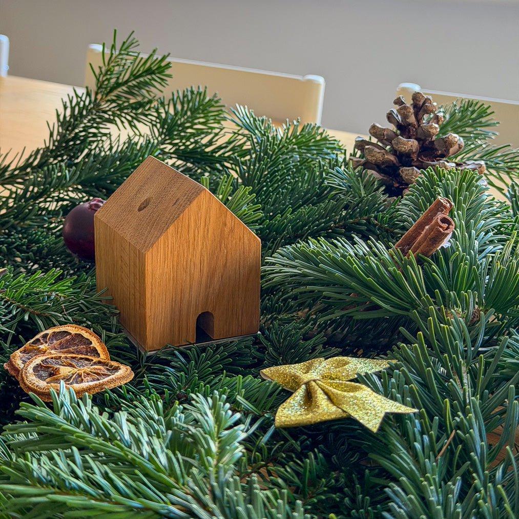 Handgefertigtes Räucherhäuschen Wieden aus recyceltem Wiener Eichenholz auf Edelstahlplatte. Platziert auf grünen Tannenzweigen und umgeben von Zimt, einer goldenen Schleife, einem Apfel, Orangenschreiben und einem Tannenzapfen.