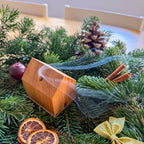Handgefertigtes Räucherhäuschen Wieden aus recyceltem Wiener Eichenholz auf einer Edelstahlplatte. Das Räucherhäuschen raucht. Platziert auf grünen Tannenzweigen und umgeben von Zimt, einer goldenen Schleife, einem Apfel, Orangenschreiben und einem Tannenzapfen.