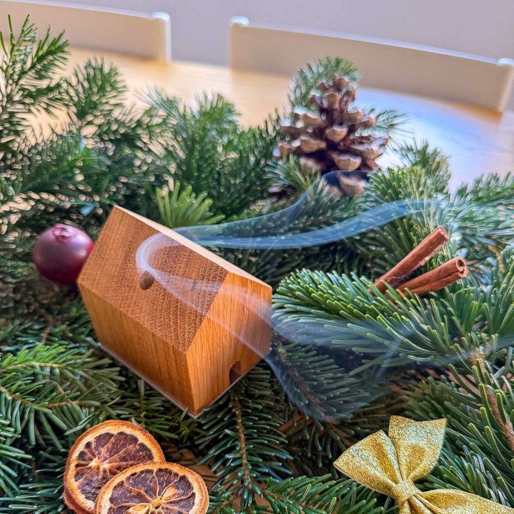 Handgefertigtes Räucherhäuschen Wieden aus recyceltem Wiener Eichenholz auf einer Edelstahlplatte. Das Räucherhäuschen raucht. Platziert auf grünen Tannenzweigen und umgeben von Zimt, einer goldenen Schleife, einem Apfel, Orangenschreiben und einem Tannenzapfen.