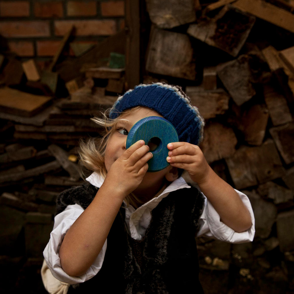 Kind spielt mit Holz Stapelturm und hält eine farbige Holzscheibe vor das Auge, Montessori Spielzeug aus Naturholz im Alltagseinsatz