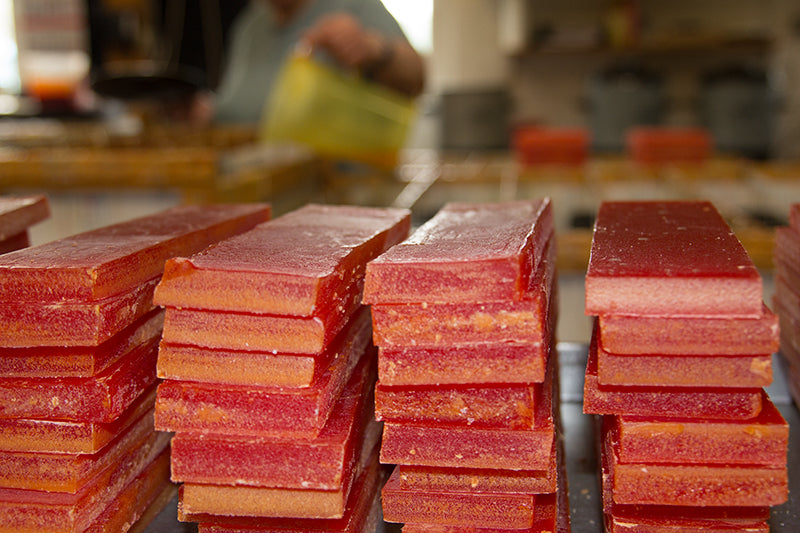 Rote Wachsblöcke im Vordergrund gestapelt. Im Hintergrund werden Kerzen von Hand gegossen. Kerzenfarm Hahn.