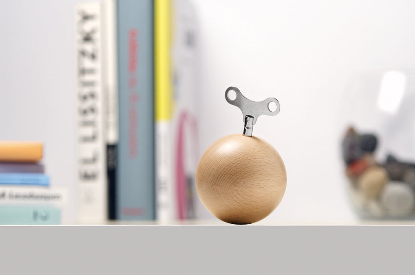 Spieluhr Mozartkugel von Siebensachen aus hellem Buchenholz mit Metallschlüssel, dekorativ auf Regal vor unscharfem Hintergrund mit Büchern