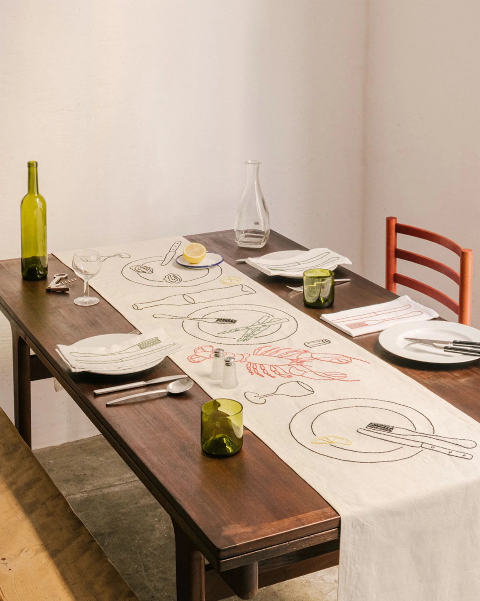 Tischläufer LE CHEMIN DU BISTROT von ŒUVRES SENSIBLES auf gedecktem Holztisch. Handgestickt auf naturfarbenem Leinen mit Motiven wie Hummer, Zitronenscheibe, Wein und Spargel. Bild ©Maxime Verret