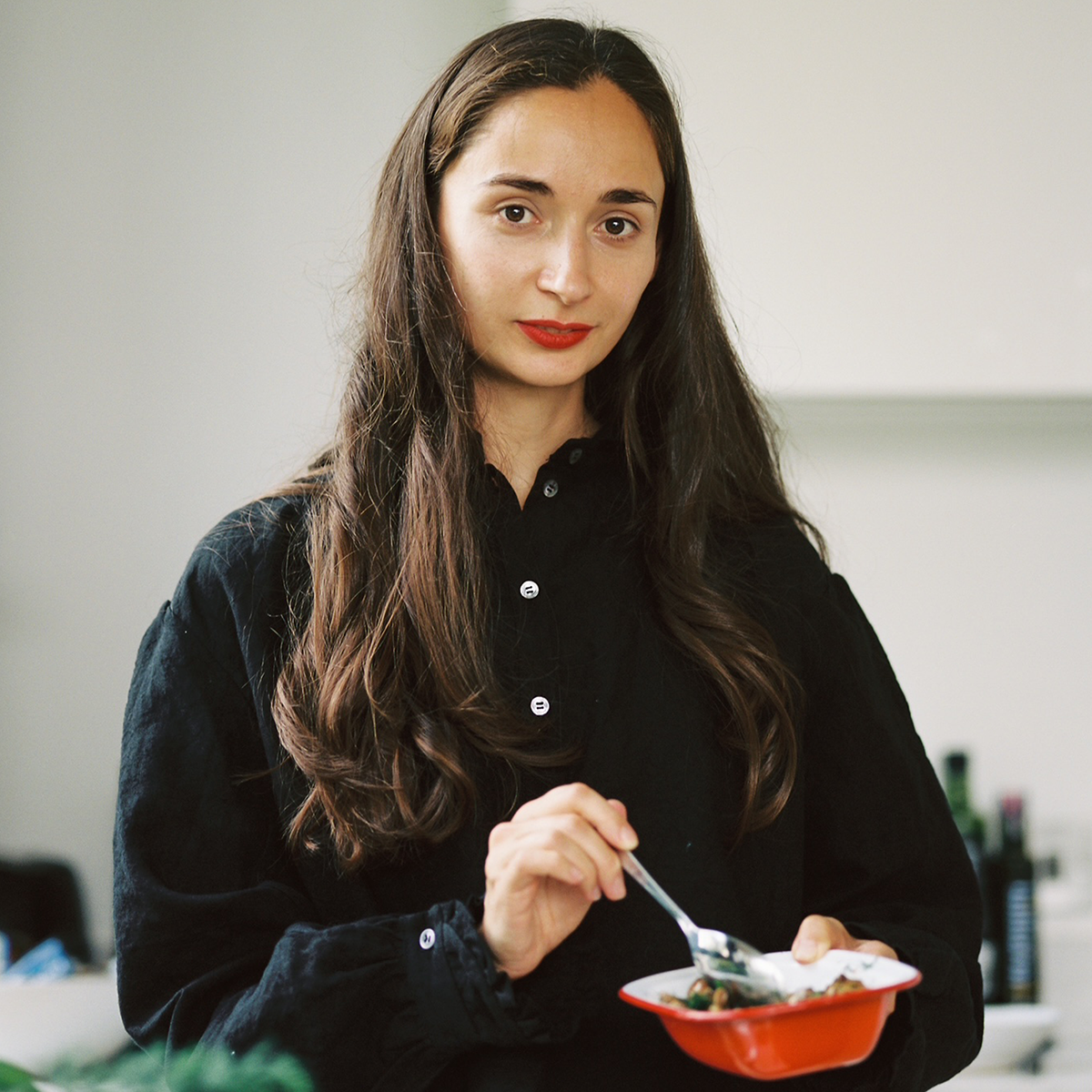 Porträtfoto von Alissa Timoshkina. Die Autorin steht in einer hellen Küche, trägt eine schwarze Bluse und hält ein rotes Schälchen mit einem Löffel in der Hand.