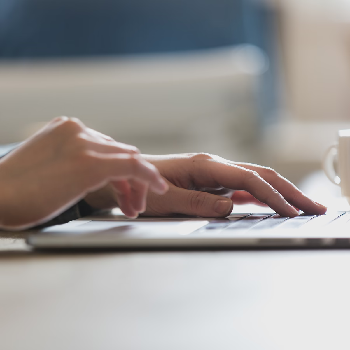 Hände einer Person tippen auf einer Laptoptastatur, im Hintergrund steht eine weiße Kaffeetasse.