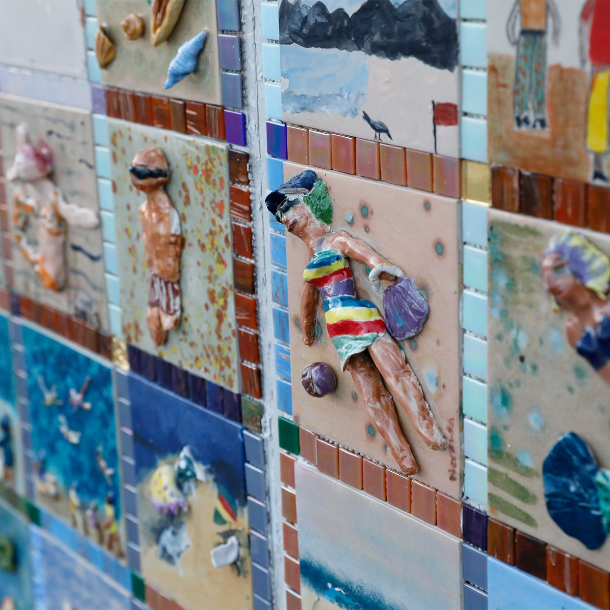 Wand aus farbigen Keramikkacheln mit plastischen Strandmotiven, darunter Figuren in Badebekleidung, eine Meerjungfrau und Strandlandschaften. Die Kacheln sind mosaikartig angeordnet und in hellen Sommerfarben gehalten.