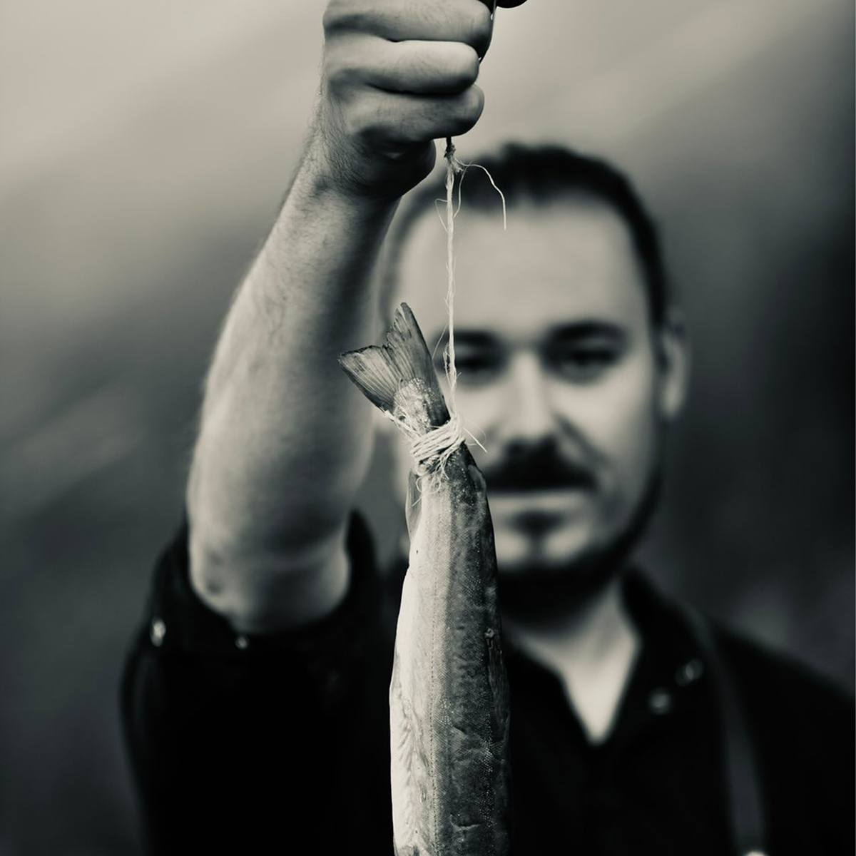 Porträtfoto von Christoph Krabichler. Der Autor hält einen Fisch an einer Schnur in die Kamera, der Fokus liegt auf dem Fisch im Vordergrund, während Krabichler unscharf im Hintergrund zu sehen ist. Schwarz-Weiß-Aufnahme.