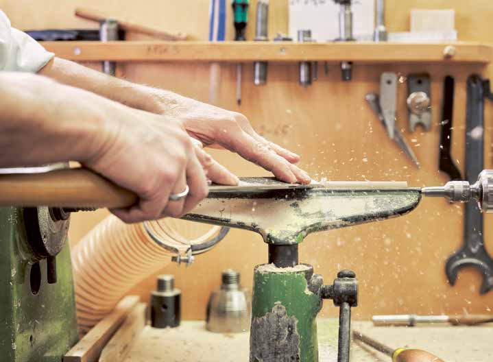Nahaufnahme der Handarbeit in der e+m Holzmanufaktur. Ein Handwerker formt ein Holzstück auf der Drechselbank während feine Späne fliegen umgeben von Werkzeugen und präziser Handwerksatmosphäre.