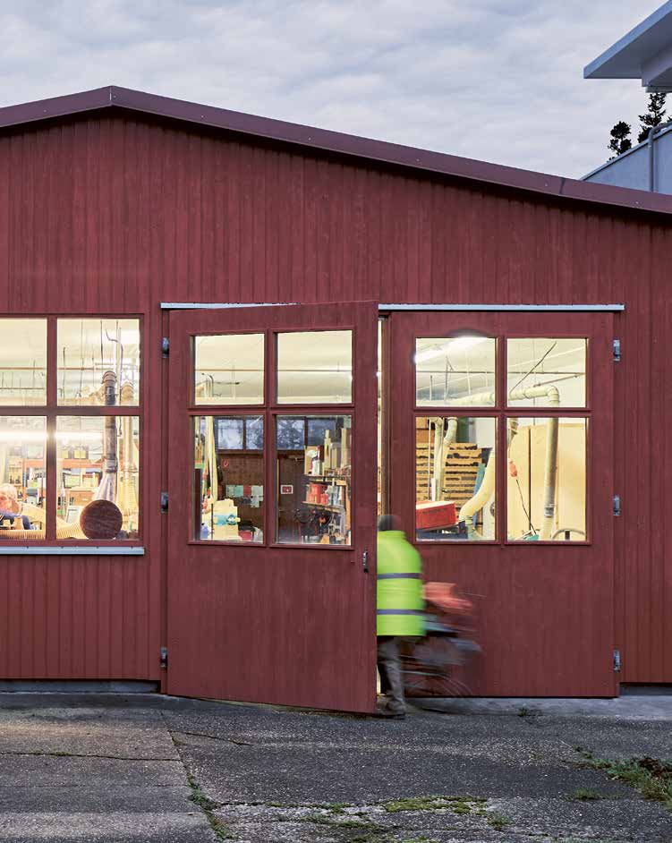 Außenansicht der e+m Holzmanufaktur in Neumarkt in der Oberpfalz. Durch die großen Fenster der rot gestrichenen Werkhalle sind Werkbänke und Maschinen zu sehen, während eine Person mit Warnweste gerade in das Gebäude geht.