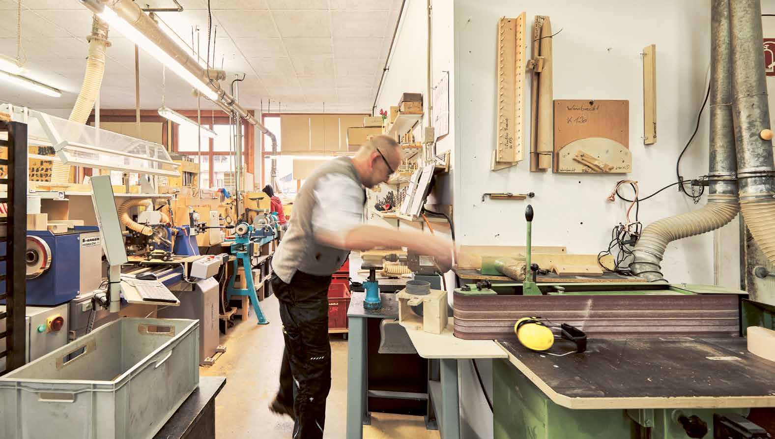 Einblick in die Werkstatt der e+m Holzmanufaktur in Neumarkt in der Oberpfalz. Ein Handwerker arbeitet konzentriert an einer Schleifmaschine, umgeben von Werkzeugen, Holzmaterialien und Maschinen. Ein authentischer Moment traditioneller Handwerkskunst.