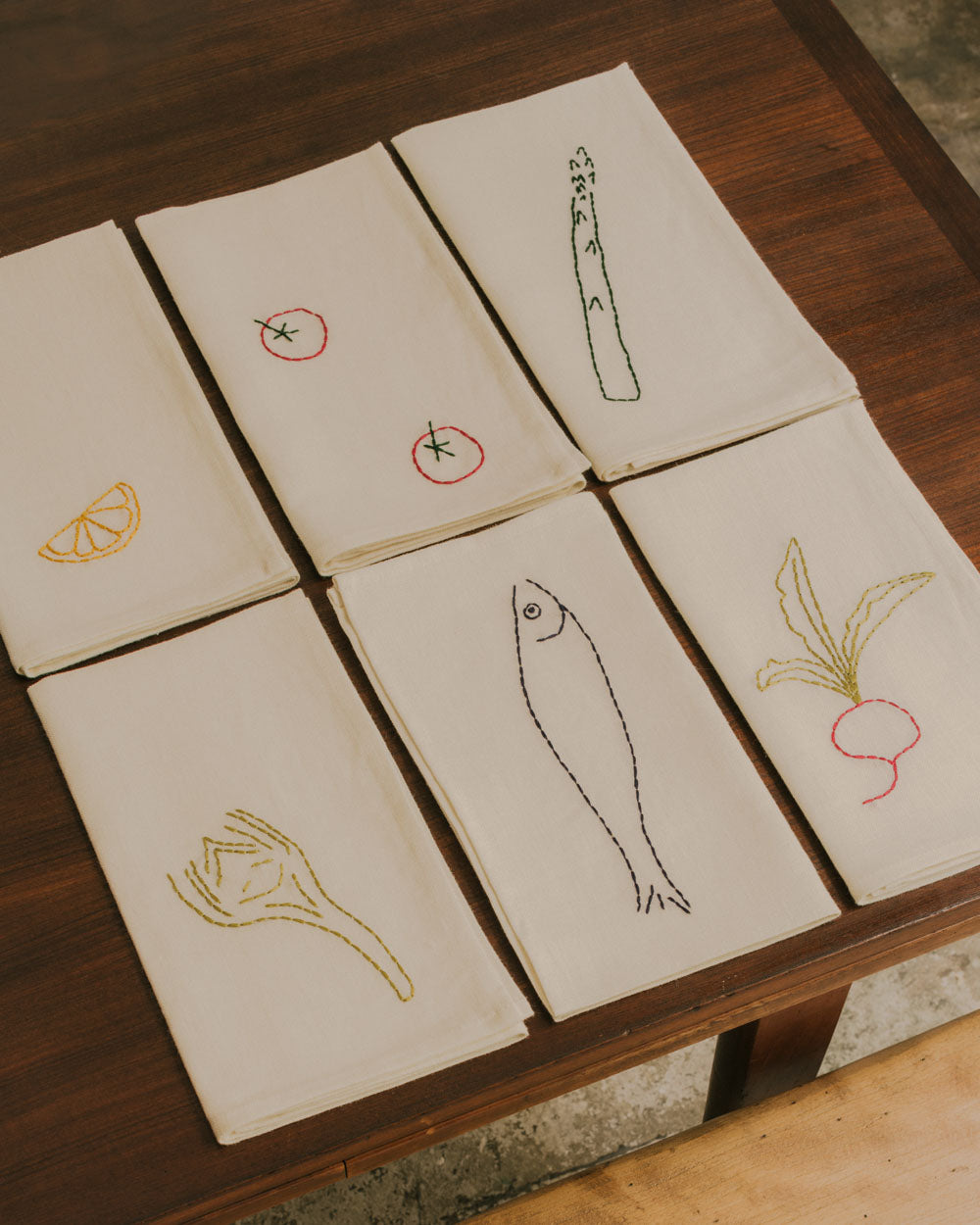 Handgestickte Servietten DINER AU BISTROT von ŒUVRES SENSIBLES aus 100 % Leinen. Sechs Motive – Spargel, Zitrone, Fisch, Radieschen, Tomaten und Fenchel auf Holztisch arrangiert. Bild © Maxime Verret