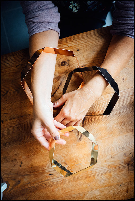 Zwei Hände halten die drei Elemente des Design-Untersetzer-Sets HEXAGON von Siebensachen in den Farben Messing, Kupfer und Gold. Die polierten Metallbänder zeigen ihre glänzende, prismatische Form auf einer Holzoberfläche. Teil der A+H Foreign Affairs Reihe.