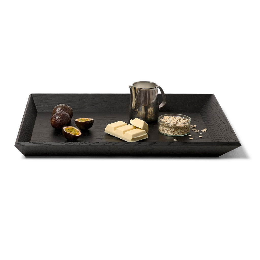 Black wooden tray OBLIQUE by NIEVEN with slanted edges. Arranged on top: passion fruits, white chocolate, a milk jug and a glass of oats. A modern minimalist serving tray with architectural design by Jörg Adam.