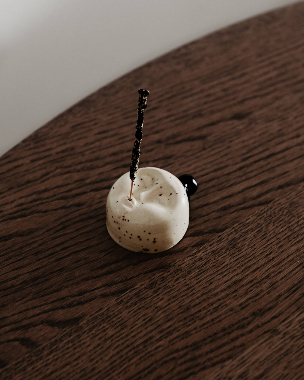 Minimalistischer Keramik-Räucherstäbchenhalter in Gesichtsform mit brennendem Räucherstäbchen auf einem Holztisch. Handgefertigtes Designobjekt aus cremefarbener Glasur mit dunklen Sprenkeln, geschaffen für ruhige Momente und modernes Interior.