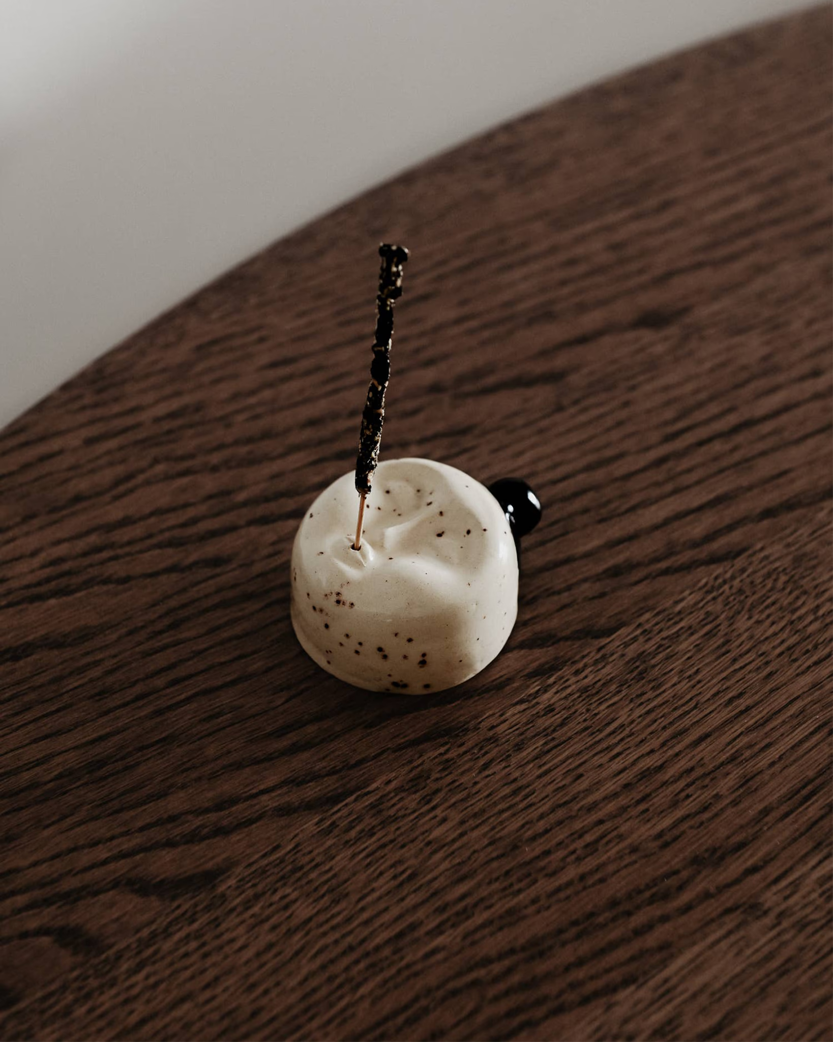 Minimalistischer Keramik-Räucherstäbchenhalter in Gesichtsform mit brennendem Räucherstäbchen auf einem Holztisch. Handgefertigtes Designobjekt aus cremefarbener Glasur mit dunklen Sprenkeln, geschaffen für ruhige Momente und modernes Interior.