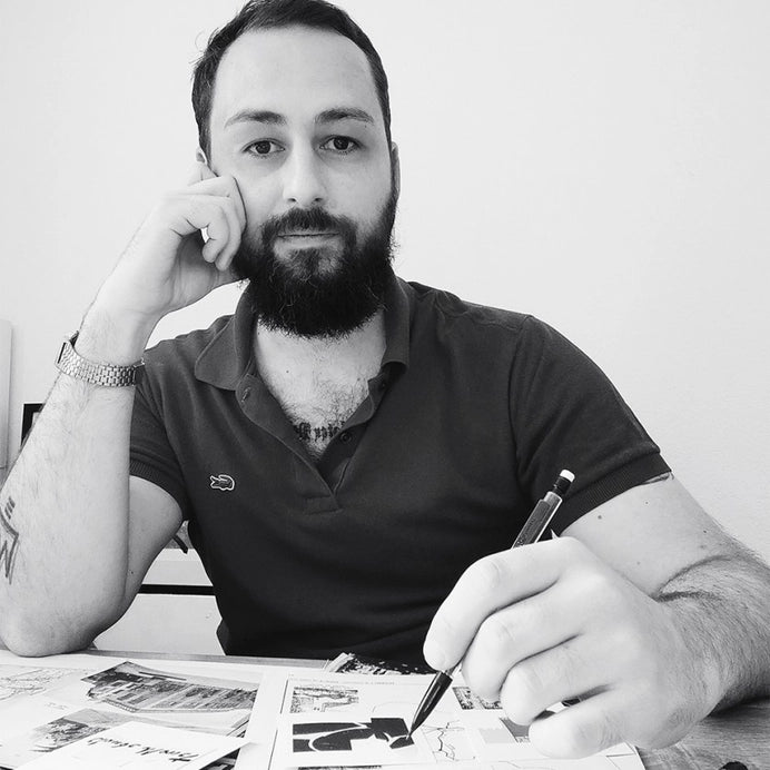 Porträtfoto von Jean-Philippe Henric. Der Künstler sitzt an einem Tisch mit Zeichnungen und hält einen Stift in der Hand, trägt ein dunkles Poloshirt und blickt ruhig in die Kamera.