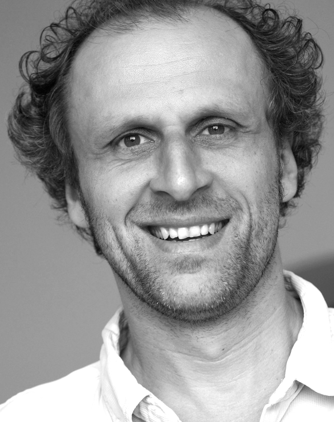 Porträtfoto von Jörg Adam. Der Designer und Gründer von NIEVEN lächelt in die Kamera, trägt ein helles Hemd und steht vor neutralem Hintergrund.