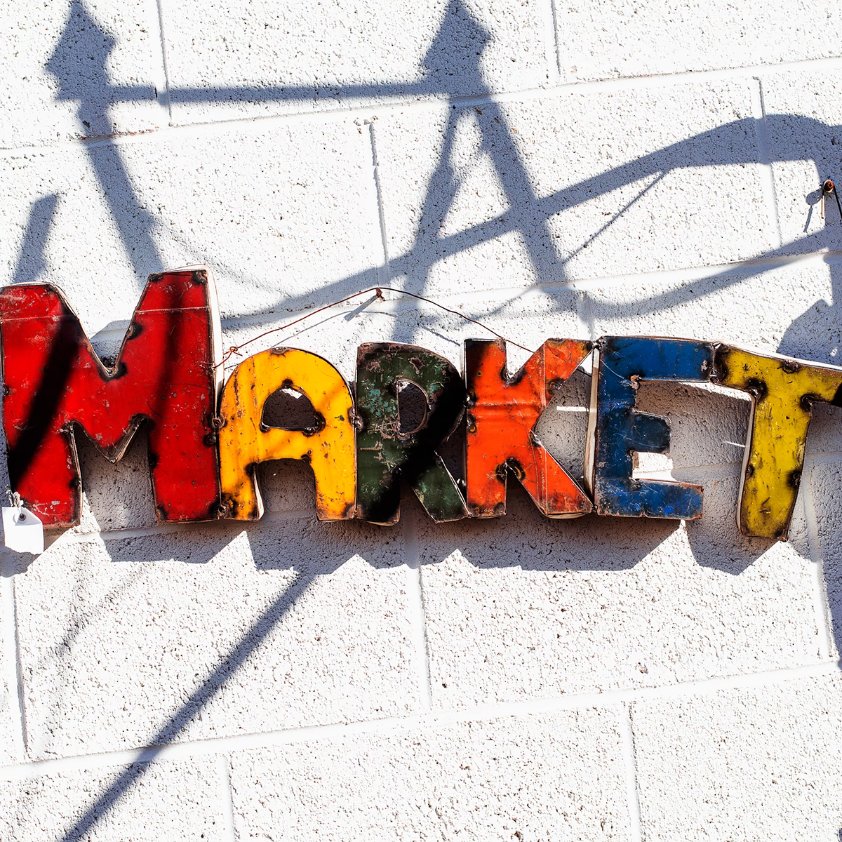 Bunte Metallbuchstaben an einer weißen Wand formen das Wort ‚Market‘. Die Buchstaben wirken gebraucht und sind in Rot, Gelb, Grün und Blau bemalt.