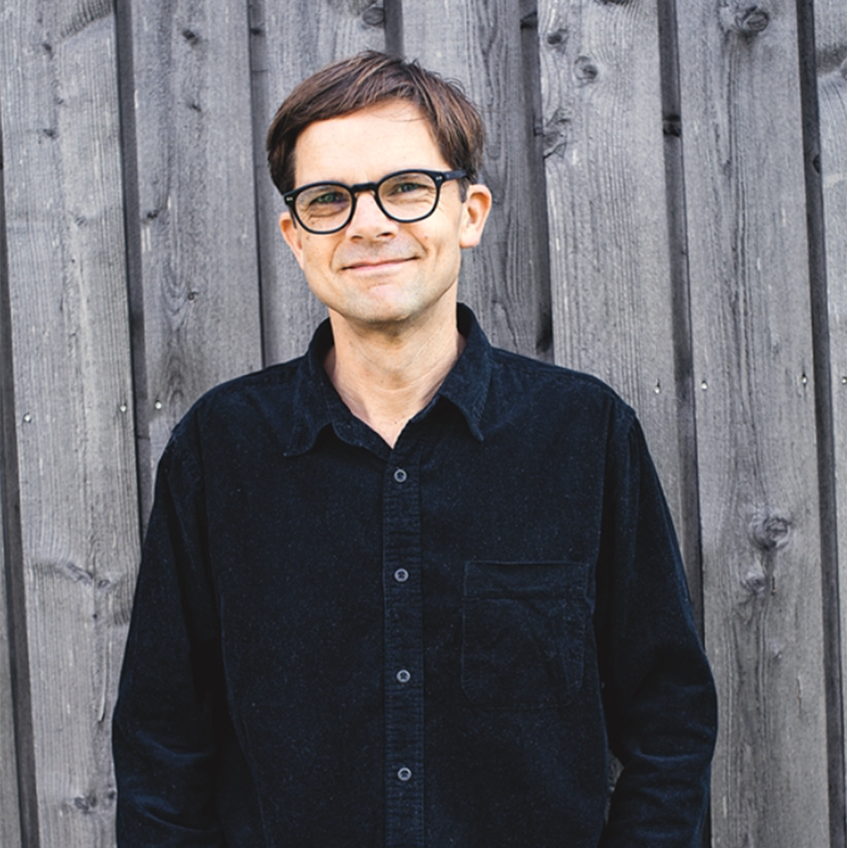 Porträtfoto von Martin Johansson. Der Autor steht lächelnd vor einer Holzwand und trägt ein schwarzes Hemd mit Kragen und Brusttasche.