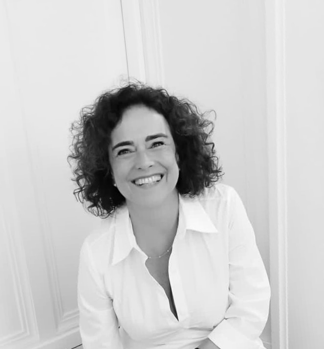 Porträtfoto von Mica Barbot, französisch-brasilianische Künstlerin und Gründerin des Atelier Raspail in Nizza. Sie lächelt in die Kamera, trägt ein weißes Hemd und sitzt vor einer hellen Wand. Schwarz-Weiß-Aufnahme.