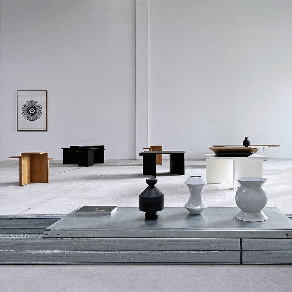 Ausstellungsansicht von NIEVEN mit minimalistischen Designobjekten und Möbeln in Schwarz, Weiß und Holz. Im Vordergrund stehen skulpturale Vasen, im Hintergrund moderne Beistelltische in einer lichtdurchfluteten Galerie.