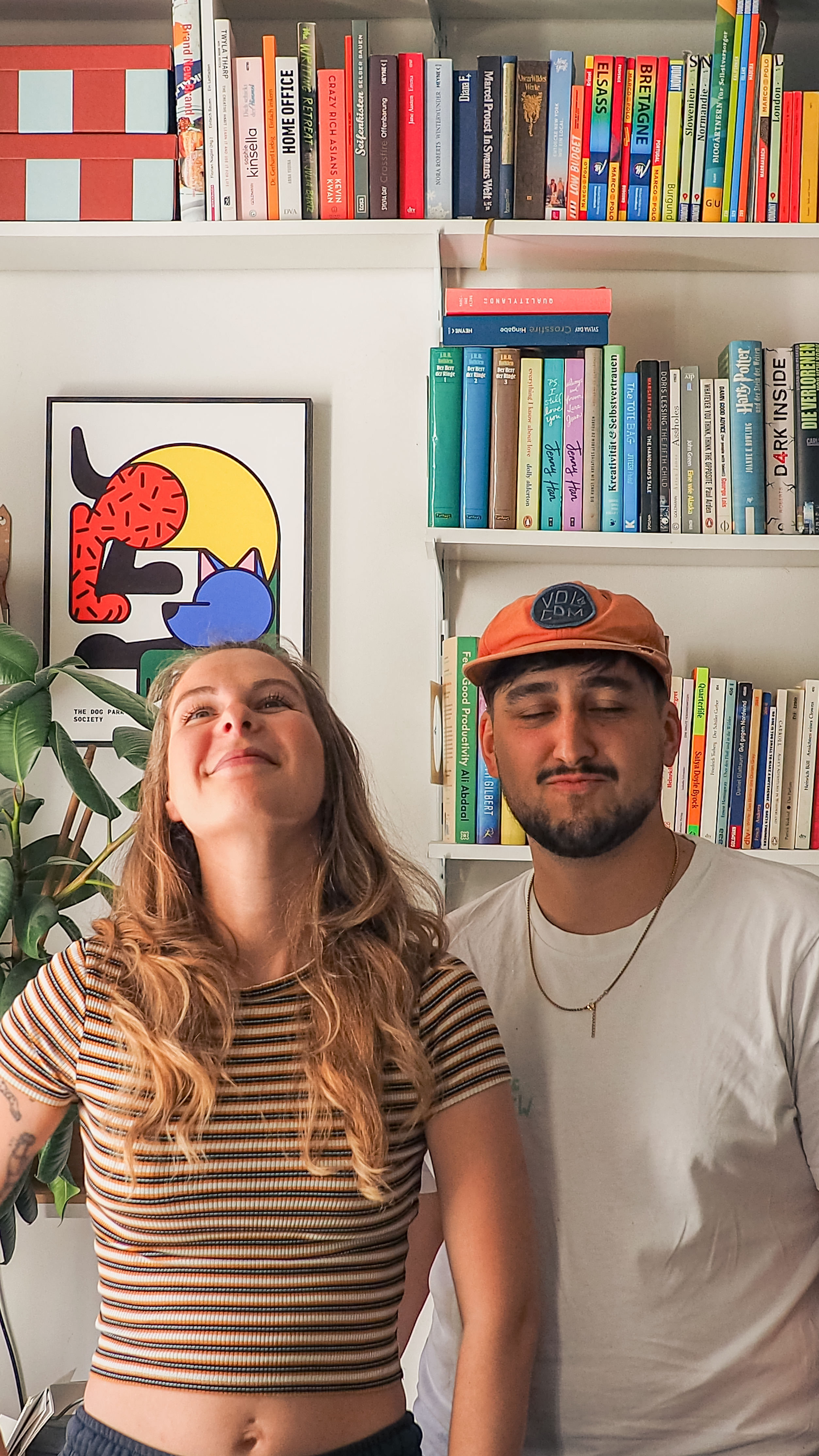 Porträtfoto des Design-Duos RadandYellow aus Wien, bestehend aus Lisa und Johann. Beide stehen lächelnd vor einem Bücherregal, sie trägt ein gestreiftes Shirt, er eine orangefarbene Cap und ein weißes T-shirt mit einer goldene Kette.