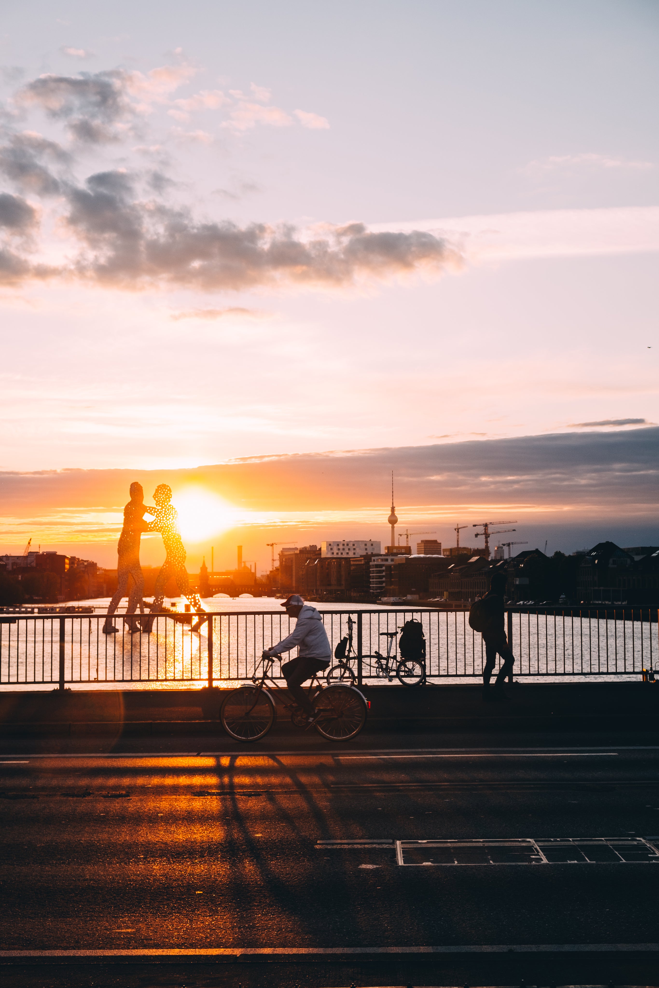 Sonnenuntergang über Berlin mit Blick auf das Molecule Man, die Spree und den Fernsehturm; Radfahrer und Fußgänger auf der Oberbaumbrücke im warmen Abendlicht.