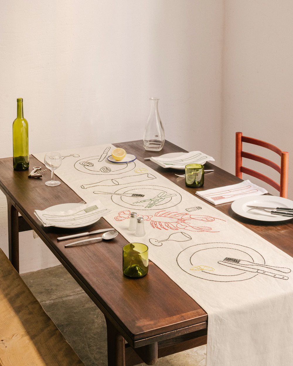 Tischläufer LE CHEMIN DU BISTROT von ŒUVRES SENSIBLES auf gedecktem Holztisch. Handgestickt auf naturfarbenem Leinen mit Motiven wie Hummer, Zitronenscheibe, Wein und Spargel. Bild ©Maxime Verret