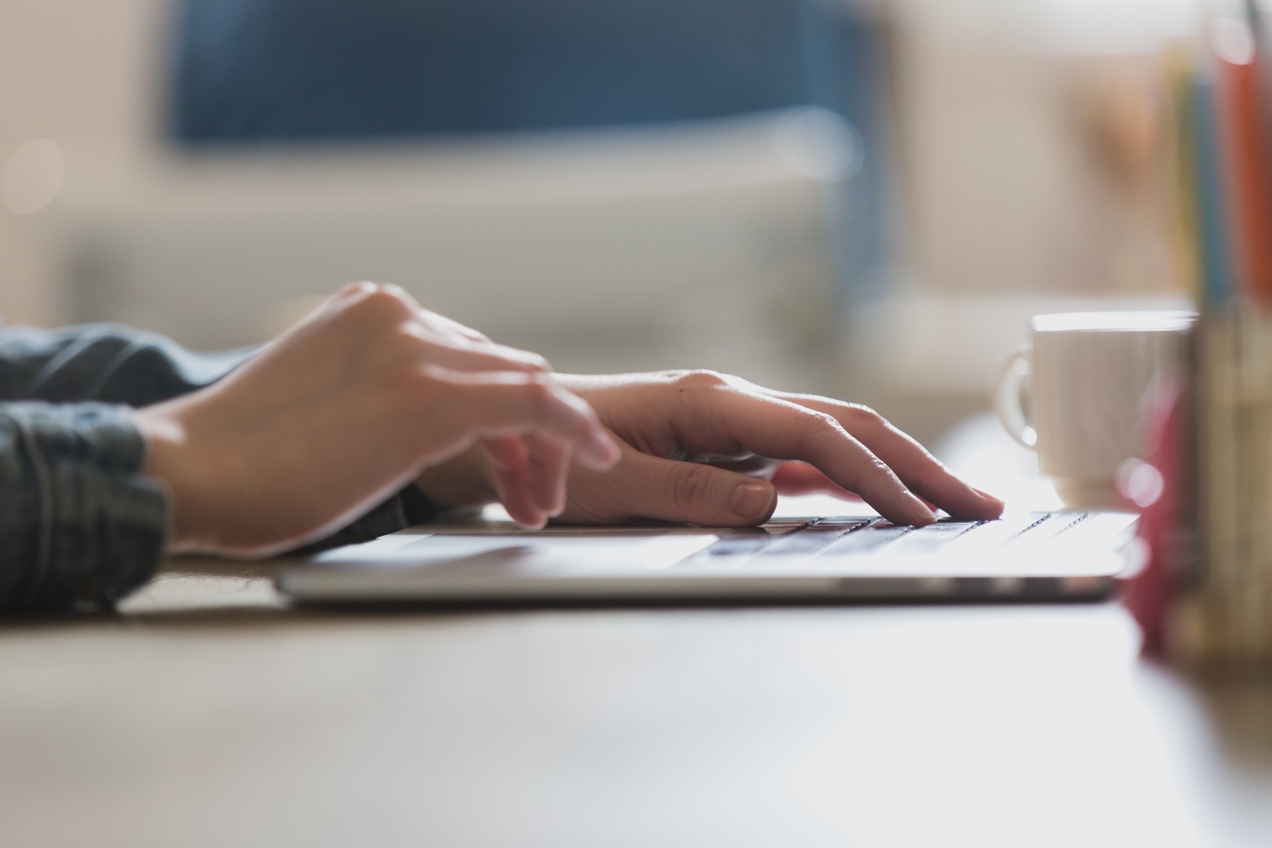 Hände einer Person tippen auf einer Laptoptastatur, im Hintergrund steht eine weiße Kaffeetasse.