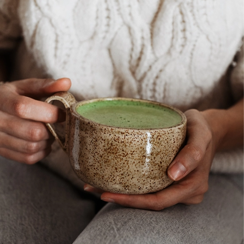 Handgefertigte Keramiktasse Caliente von Üm Studio in warmem Beige mit feinen braunen Sprenkeln, gehalten in beiden Händen. Gefüllt mit cremigem Matcha Latte, ein Moment der Ruhe und Achtsamkeit, der die Wärme Málagas in den Alltag bringt.
